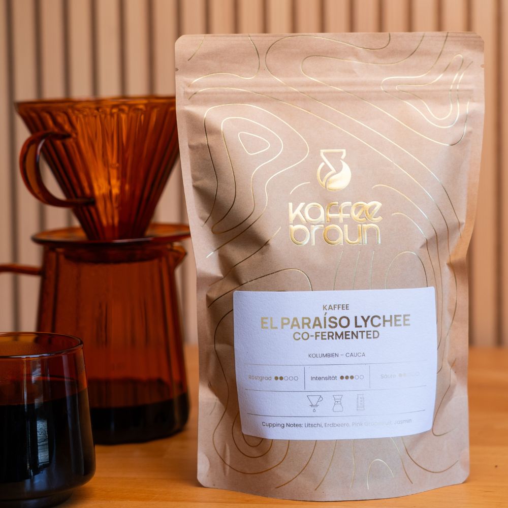 Packung El Paraíso Lychee steht neben einer Filterkanne aus braunem Glas mit frisch gebrühtem Kaffee