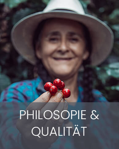 Philosophie & Qualität Eine Kaffeefarmerin hält lächelnd rote Kaffeekirsche in die Kamera, das Bild ist halb geteilt und die untere Hälfte blau hinterlegt mit der Aufschrift Philosophie & Qualität