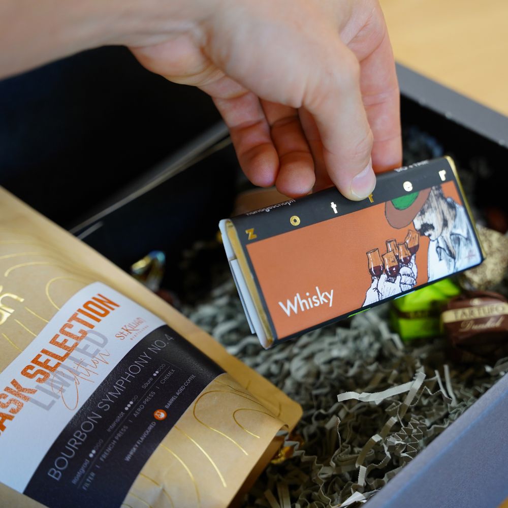 Hand hält Zotter Whisky-Schokolade über der Geschenkbox, daneben der Kaffee Braun Cask Selection Beutel und Pralinen