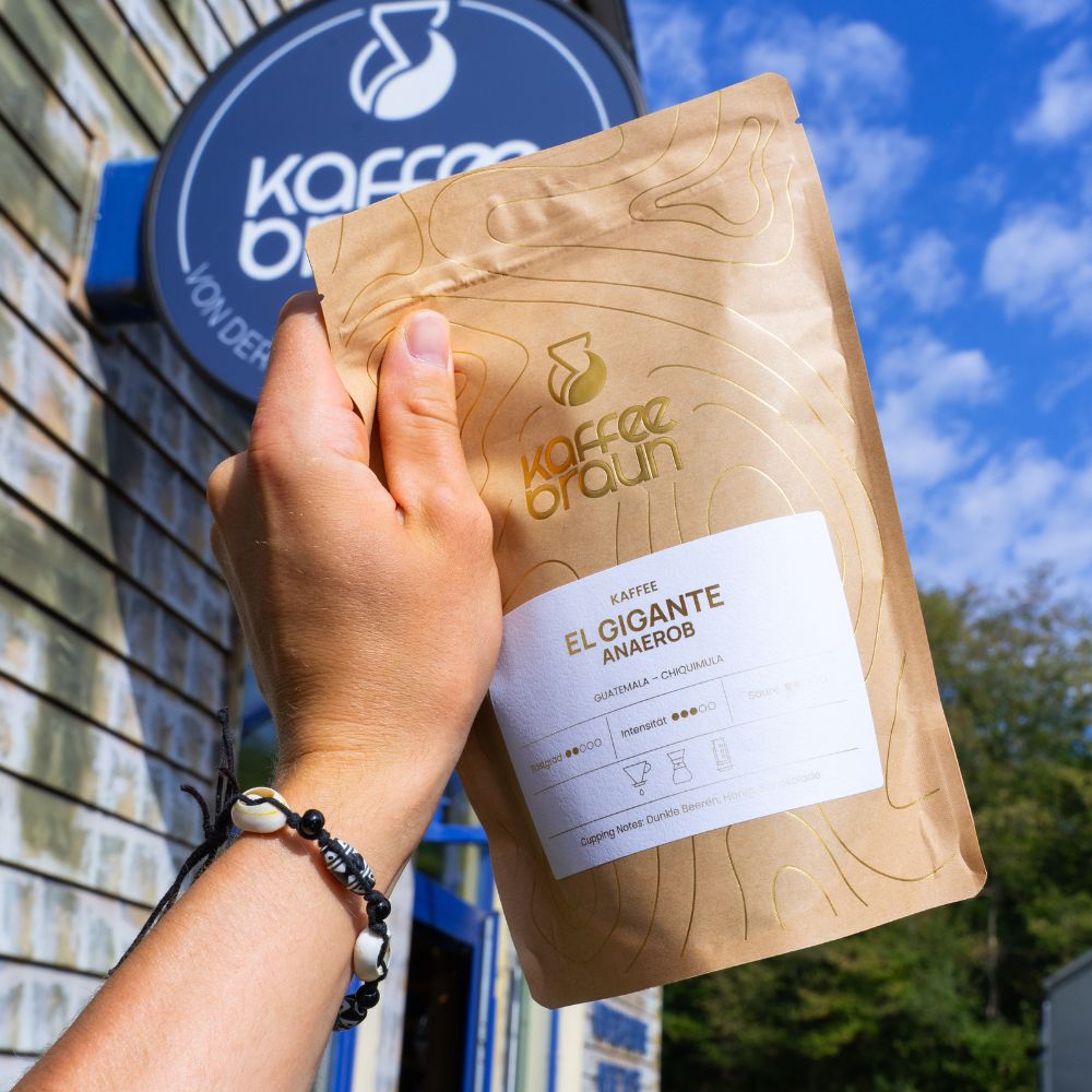 Hand hält eine Packung Kaffee Braun El Gigante Anaerob vor dem Rösterei-Schild, blauer Himmel im Hintergrund