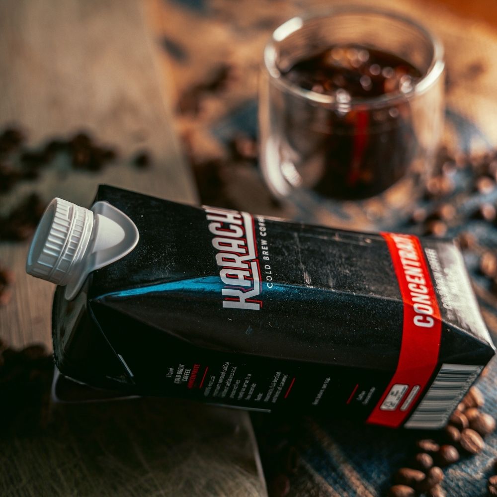 Schwarzer TetraPak KARACHO Cold Brew Coffee Concentrate neben einem Glas Cold Brew mit Eis"