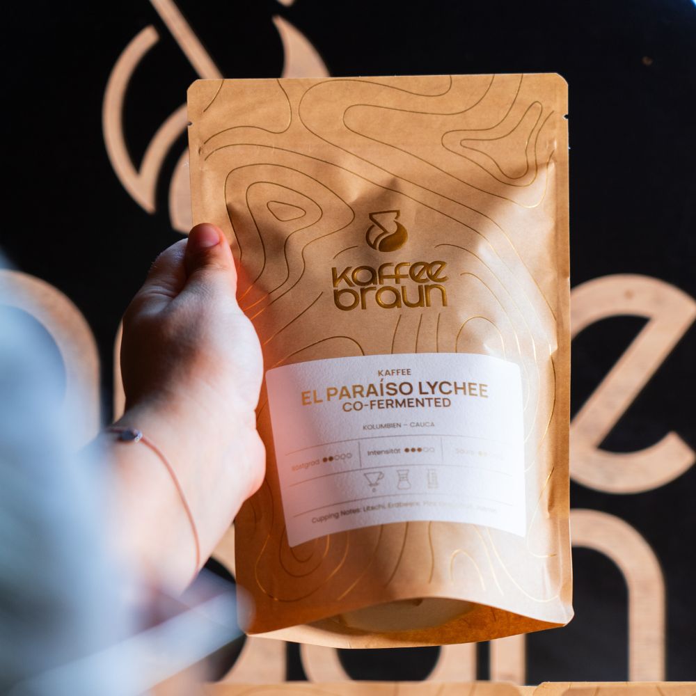 Hand hält eine Packung El Paraíso Lychee vor dunklem Hintergrund mit Kaffee-Braun-Logomuster